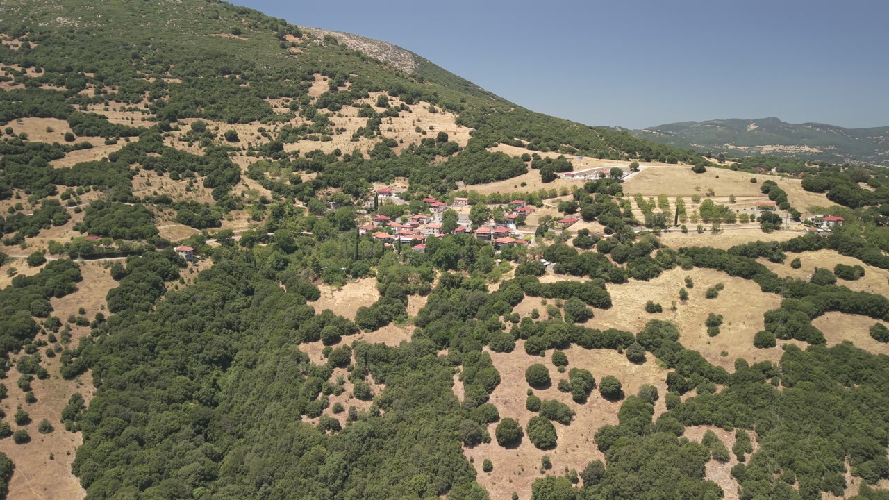 Aerial drone pull back reveal shot of a traditional Greek village in mikros pontias, surrounded by scenic mountain landscape on a sunny day.
