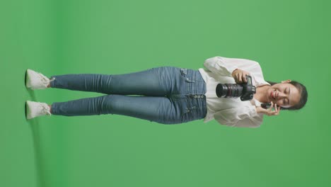cuerpo lleno de fotógrafo asiático sosteniendo una cámara en sus manos y hablando en teléfono inteligente mientras está de pie en el fondo de la pantalla verde en el estudio