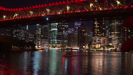 Time-lapse shot of a dazzling cityscape at night showcases a red-lit Story bridge over Brisbane river with illuminated skyscrapers reflecting off the water, creating a vibrant urban scene.