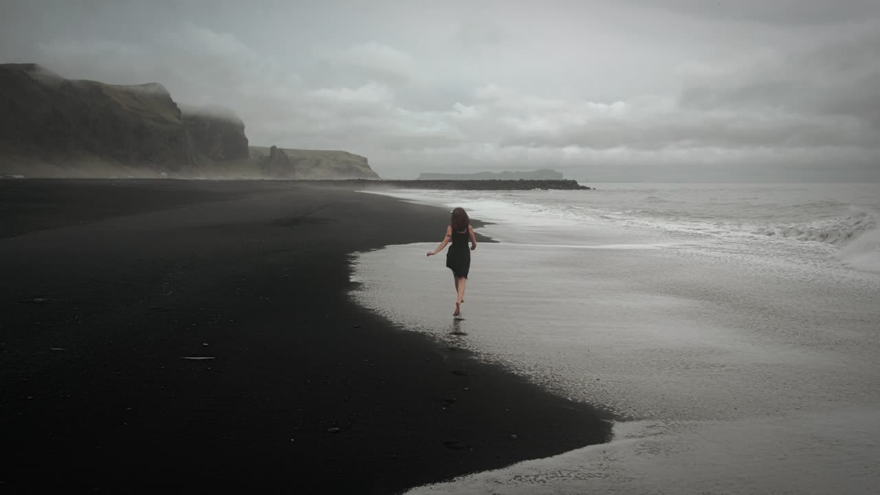 joven hermosa con vestido negro corriendo en la playa de arena negra de islandia a través de las olas, disparo de seguimiento aéreo