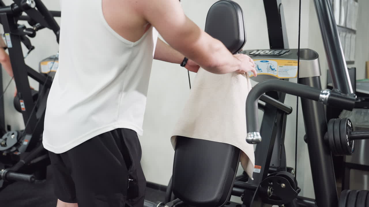Man spreads gym towel on chair seat before workout, preparing bench with towel draped over backrest in modern fitness center with sleek equipment and bright interior lighting