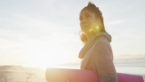 Mujer-De-Raza-Mixta-Sosteniendo-Una-Estera-De-Yoga-En-La-Playa-Sonriendo