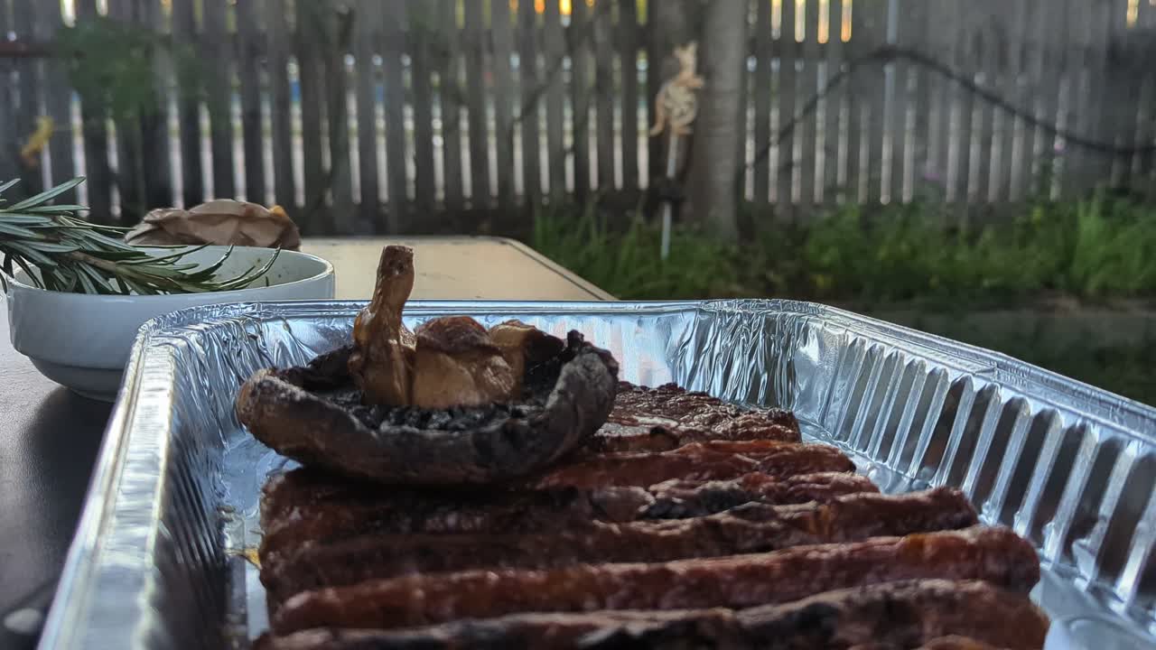 Tilt up, Barbequed sausages and grilled mushroom in a foil tray with garden fence and kangaroo feature in background.