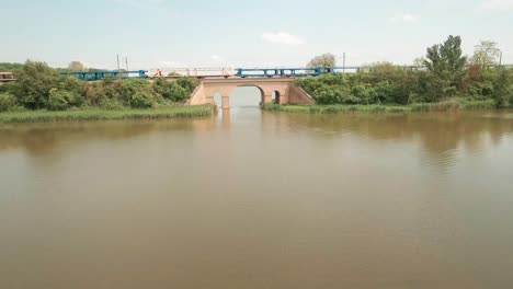 Aerial-transportation-video-of-a-drone-gliding-low-above-the-lake,-passing-through-an-opening-of-a-railway-bridge