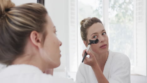 Biracial-woman-applying-beauty-face-mask-looking-at-mirror-in-bathroom,-slow-motion