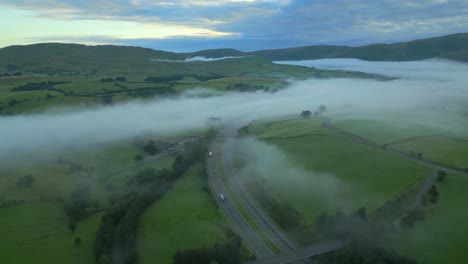 Flying-high-over-M6-motorway-with-fog-in-valley-amongst-green-rural-landscape-at-sunrise