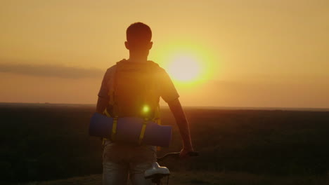 un hombre con una mochila y una bicicleta se para y mira el horizonte donde el sol se pone en bicicleta y