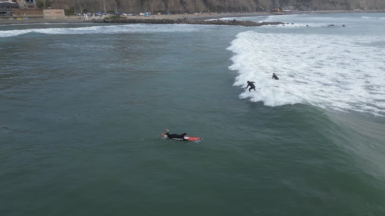 페루의 미라플로레스 리마에서 파도와 함께 바다의 공중 낮은 각도 패닝 샷, 서핑 보드에 서퍼와 바다로 수영하는 다른 사람들