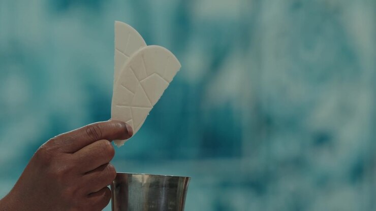 Priest hand holds white communion host above silver chalice at Catholic Mass