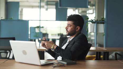 portrait-of-a-handsome,-stylish-young-man-of-Arab-descent-sitting-in-a-modern-business-center-office-with-a-laptop,-recording-a-voice-message-on-his-smartphone-while-speaking