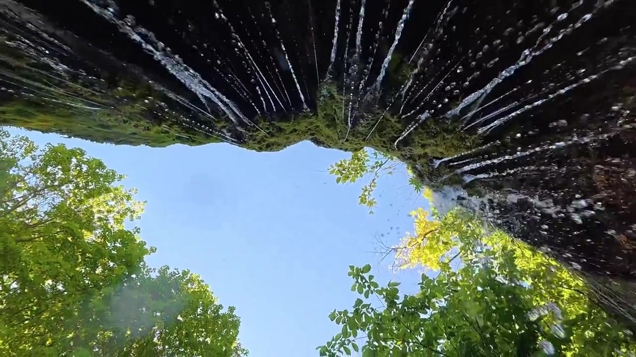 A low-angle view captures water cascading over moss-covered rocks, creating a natural curtain with a bright blue sky and green trees in the background