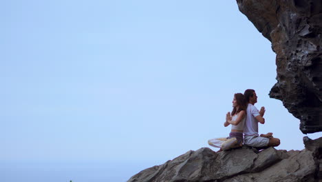 A-man-and-woman-meditate-and-practice-yoga-back-to-back-on-a-mountain's-rock,-overlooking-the-ocean's-expanse-from-the-summit