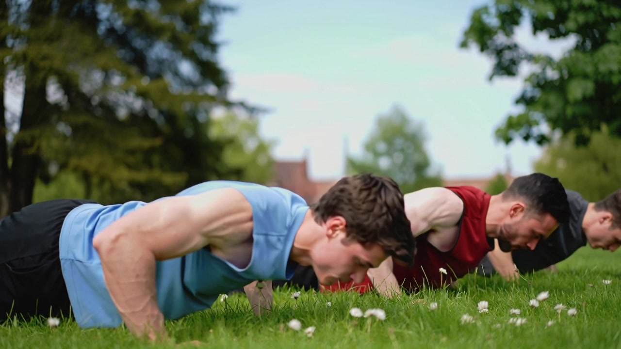 Lowering and pushing, three men training on park grass, performing push-ups in sleeveless tops
