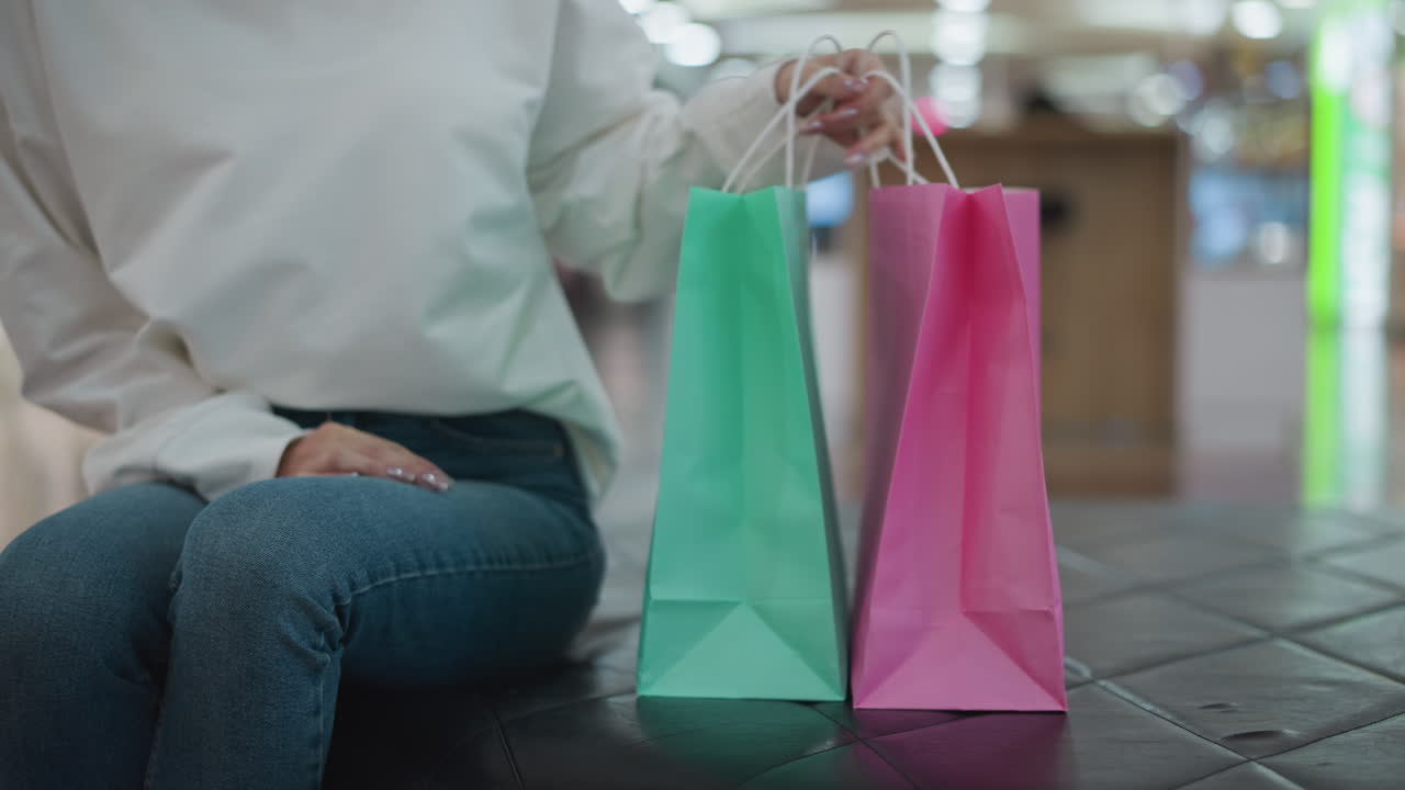 dama sentada al lado de bolsas de compras rosadas y menta en un banco de azulejos alcanza las bolsas mientras está de pie, con el fondo borroso del centro comercial con compradores y luces bokeh vibrantes