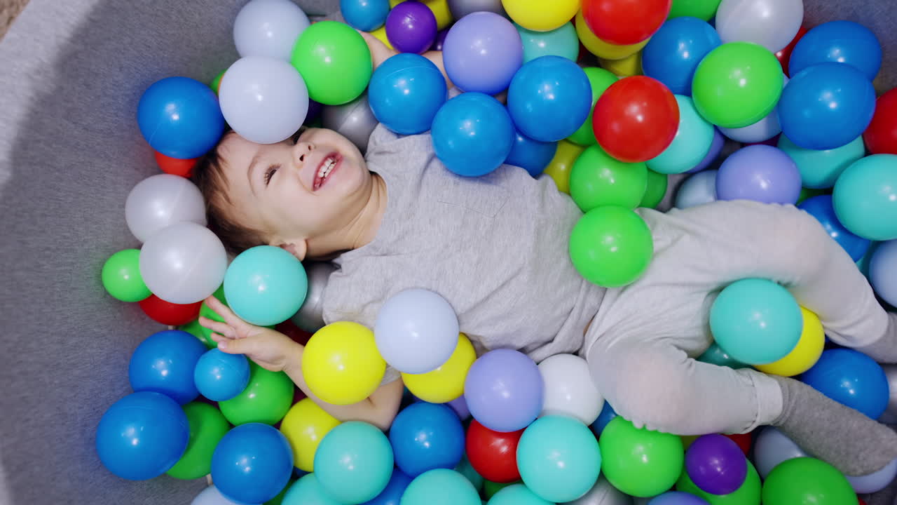 Happy smiling kid lies among the balls in dry basin. Toddler sits up us someone throws the balls to him. Top view.