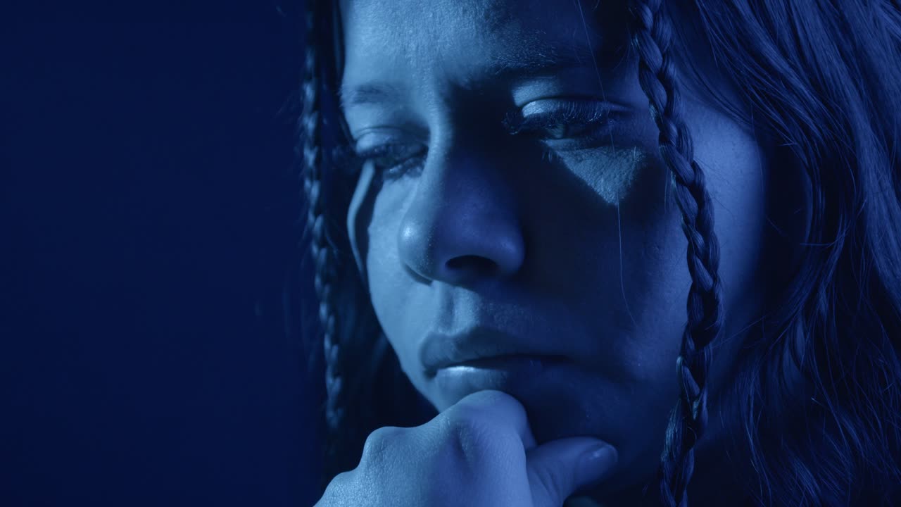 Close-up of a girl in deep reflection, face partially shadowed by moody blue lighting, adds a contemplative and introspective atmosphere. Her braided hair frames her thoughtful expression