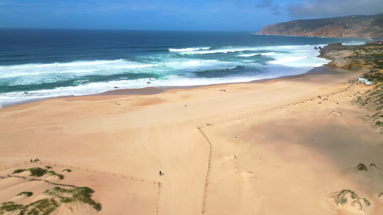 Circular aerial view of a couple walking on Guincho beach with the shadows of the clouds reflecting on the sand with the sea and the Sintra mountains in the background.Cascais,Portugal