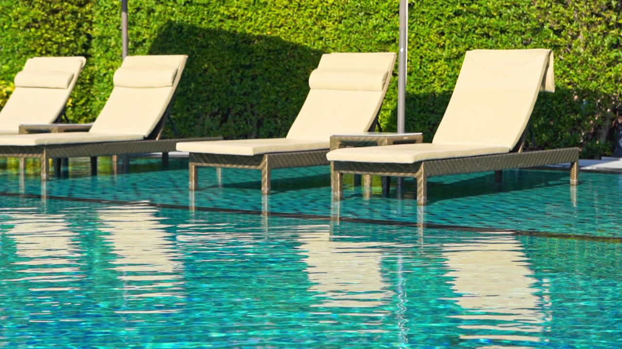 Entspannende Poolszene mit vier leeren Sonnenliegen und schimmerndem, klarem, blauem, seichtem Wasser, das die Szene und kleine Wellen widerspiegelt