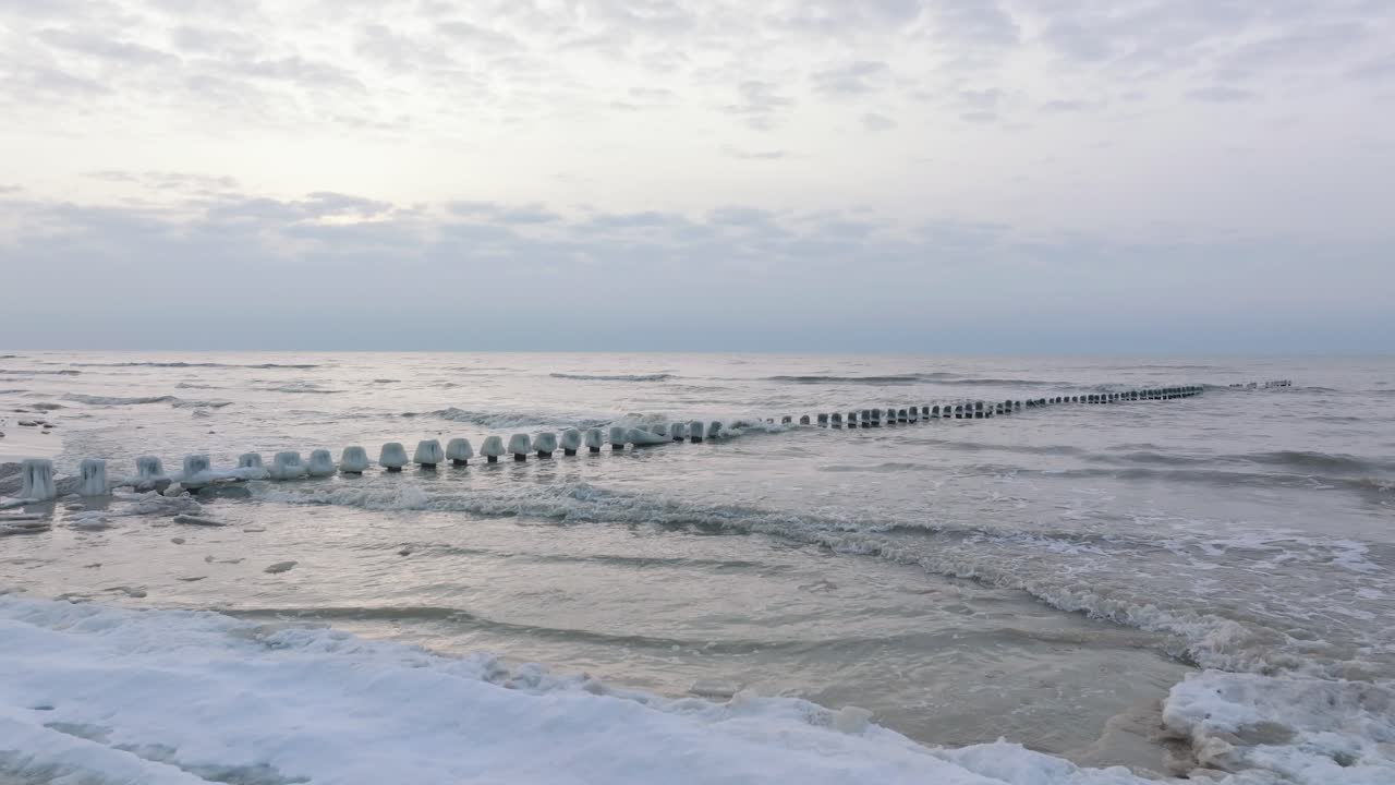 вид с воздуха на старый деревянный пирс на побережье балтийского моря, пасмурный зимний день, белый песчаный пляж, покрытый снегом, лед на деревянных столбах, спокойный берег моря, широкая съемка с дрона с низким движением вперед
