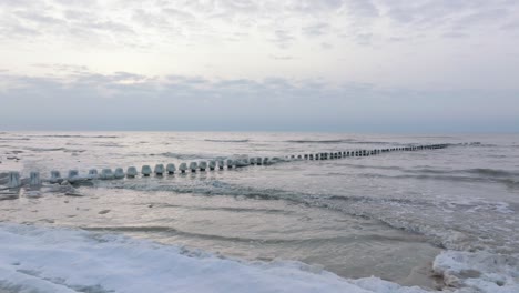 aerial establishing view of an old wooden pier at the baltic sea coastline, overcast winter day, white sand beach covered in snow, ice on wood poles, calm seashore, wide drone shot moving forward low