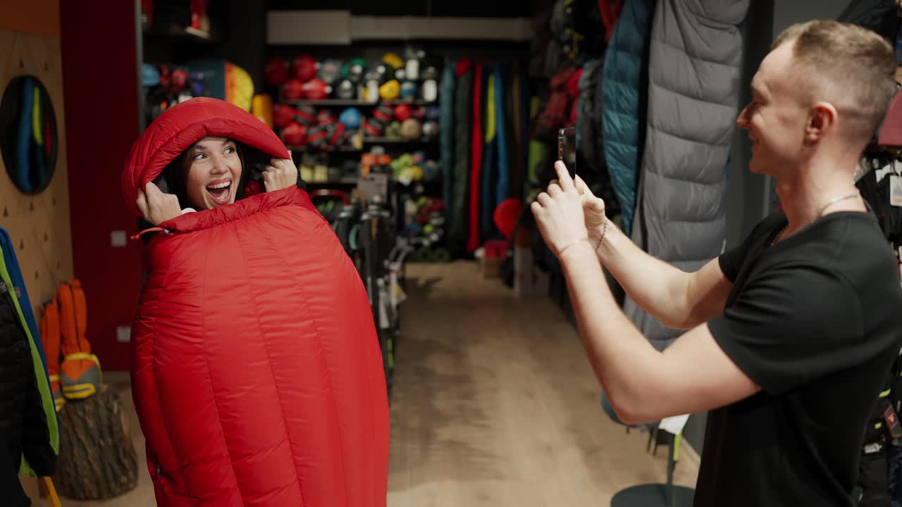una pareja sonriente comprando en una tienda de ropa deportiva tomando fotos dentro del saco de dormir