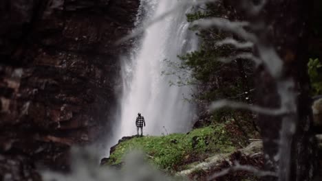 Man-standing-in-front-of-a-huge-waterfall,-slow-motion-shot-of-a-beautiful-location-during-day-time