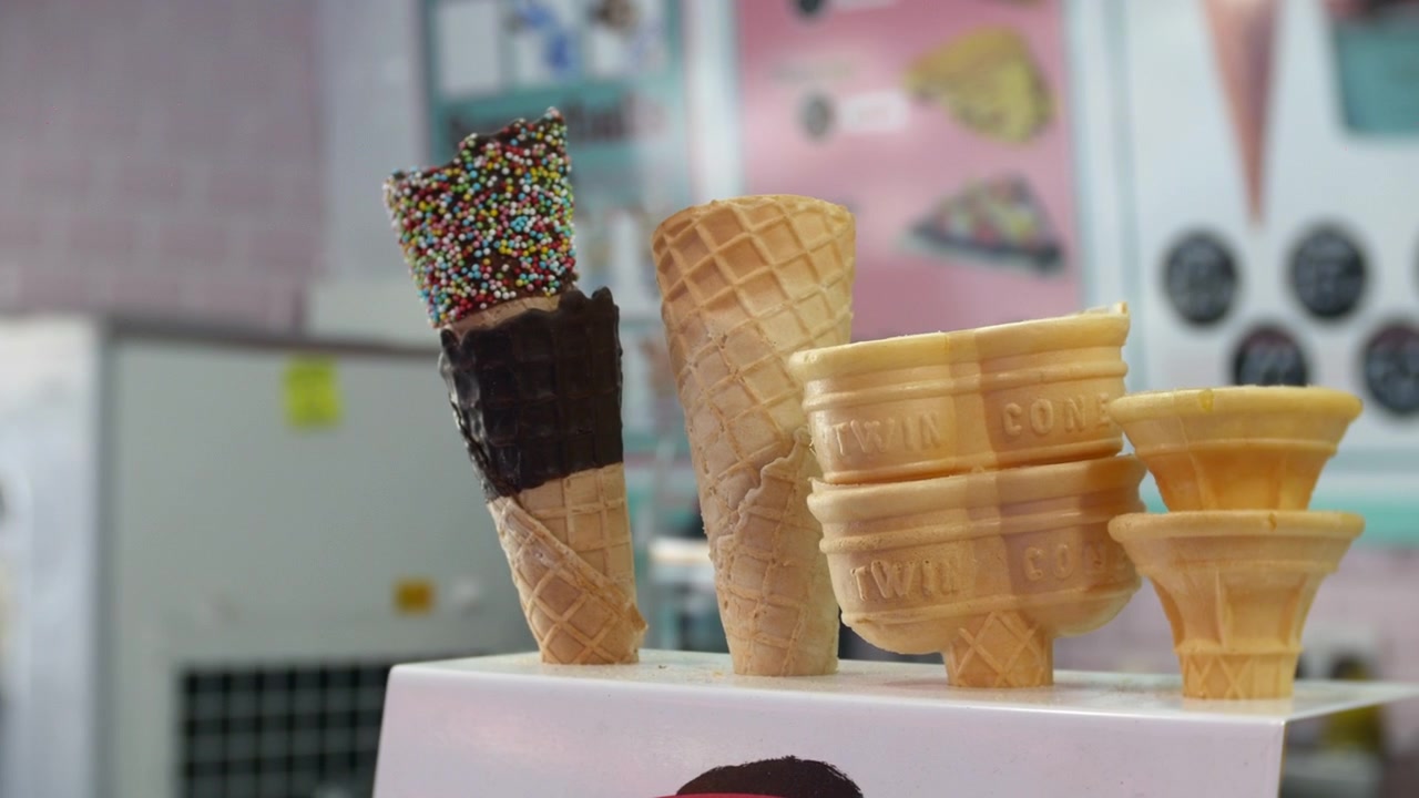 Coni disposti su un bancone includono varietà di waffle, wafer, ricoperti di cioccolato e con granella. Una vista ravvicinata evidenzia le consistenze croccanti all'interno di una gelateria dai colori pastello.