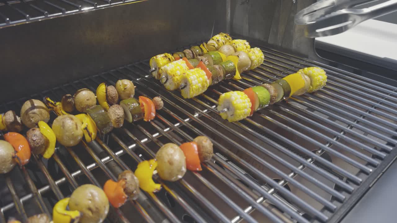 Vegetable skewers with corn, pepper, zucchini, potatoes and mushrooms are being cooked on a stainless steel bbq adjusted with stainless steel tongs