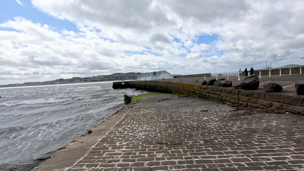 Camera pans across Broughty Ferry’s waterfront, revealing cobblestone slipway, choppy water, and historic castle under bright, partly cloudy daylight