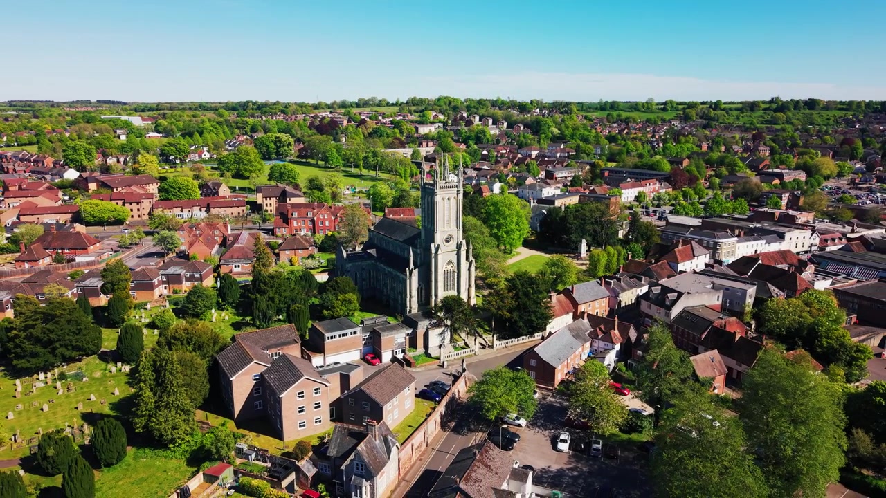 Luchtdronebeelden die terugtrekken van de St Mary's Kerk in Andover, die de historische kerktoren, het kerkhof, daken, wegen, groene ruimtes en het bredere landschap van de stad Hampshire onthullen op een zonnige dag