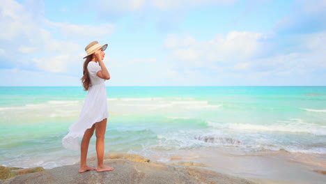 Una-Mujer-Joven-Con-Un-Vestido-De-Verano-Se-Aferra-A-Su-Sombrero-Para-El-Sol-Mientras-Mira-Las-Olas-Entrantes