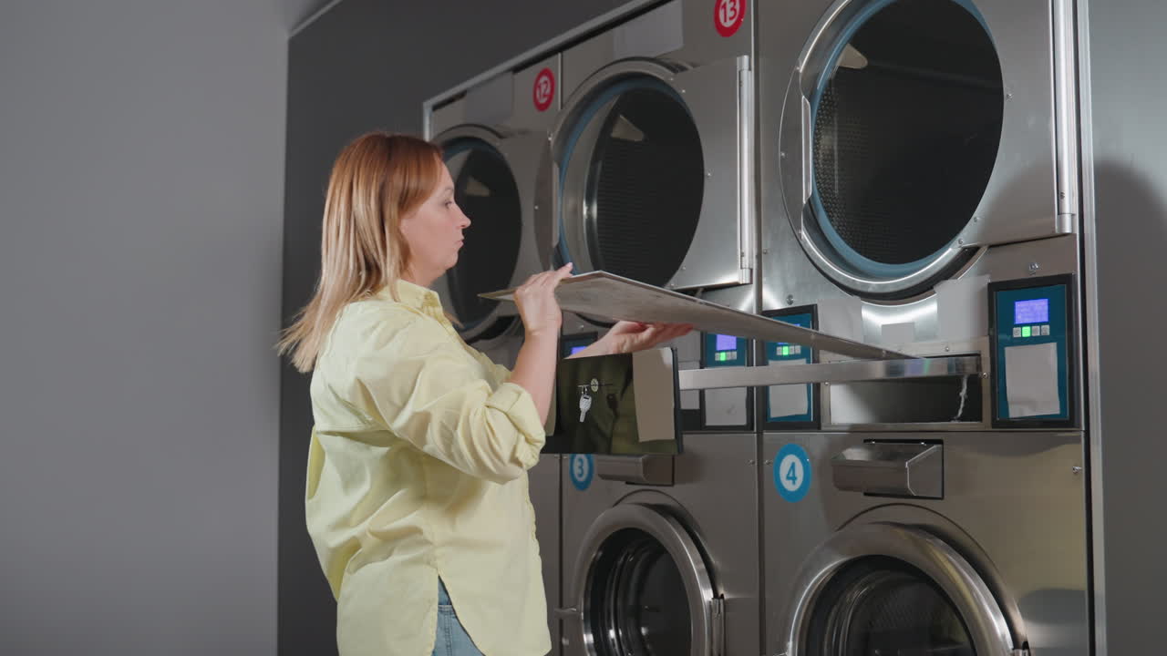 Laundress opens industrial washer filter drawer with key, removes filter, sets aside, ensures proper maintenance and hygiene inside modern laundromat, stainless machines under routine service