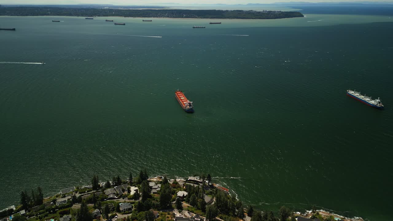 웨스트 밴쿠버의 해안선 주변을 날아다니는 드론은 푸른 바다 물, 물에 고정 된 석유 커, 산업 선박, 언덕의 초록 숲, 배경에 보운 섬을 보여줍니다.