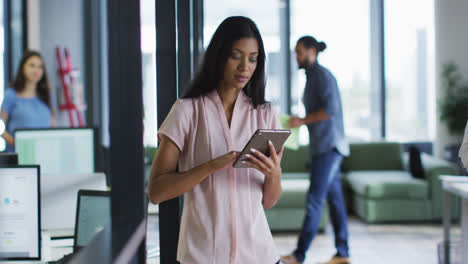 Portrait-of-mixed-race-businesswoman-standing-in-office-using-tablet-and-smiling-to-camera