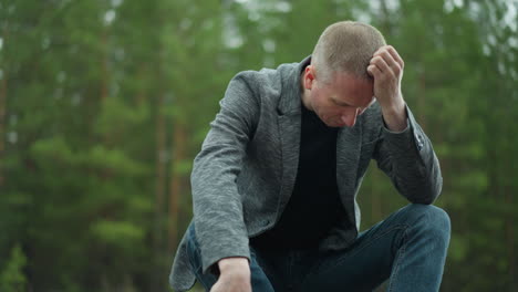 a close view of a man in a gray jacket and blue jeans sitting on a railway track, holding a handgun with his head bowed and hand resting on his head, the background shows a blurred green forest