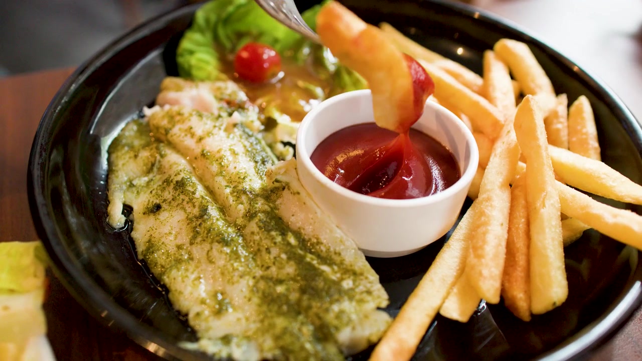 Una forchetta intinge una patatina fritta dorata nel ketchup rosso accanto a un filetto di pesce al pesto grigliato.