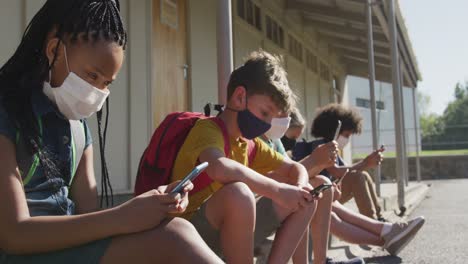 Gruppe-Von-Kindern,-Die-Gesichtsmasken-Tragen-Und-Smartphones-Benutzen,-Während-Sie-Zusammensitzen