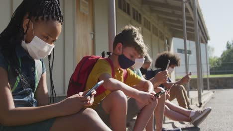 Grupo-De-Niños-Con-Máscaras-Faciales-Usando-Teléfonos-Inteligentes-Mientras-Están-Sentados-Juntos