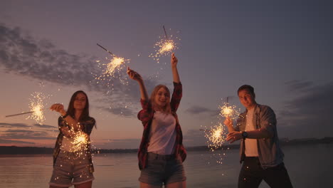 alegres amigos masculinos y femeninos están corriendo a lo largo de la playa al atardecer sosteniendo fuegos artificiales brillantes y luces fugitivas en cámara lenta. baile y fiesta de atardecer en la playa