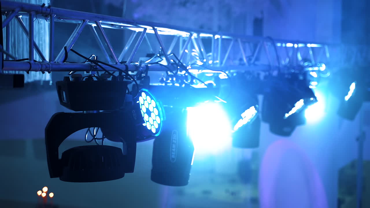 Black projectors spinning and giving bright lights in the night club. Metal construction and many spotlights hanging on it on the blue blurred background inside the hall.