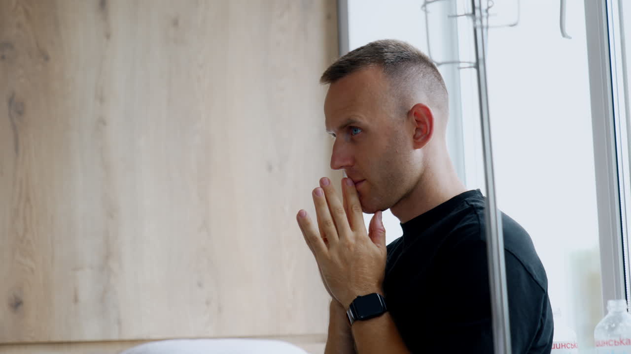 Tensed Caucasian man in black t-shirt standing at the window. Nervous man holding hands in front of his face. Husband worries for wife at childbirth.