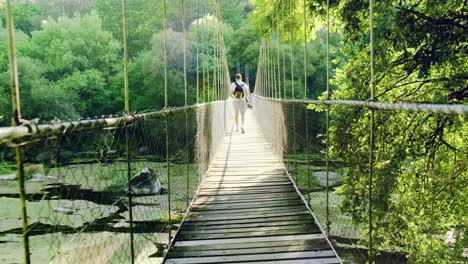 Adventurer-photographer-walk-away-from-camera,-crossing-Supension-rope-bridge-surrounded-by-lush-vegetation