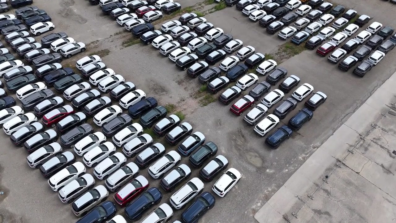 Automóviles de pasajeros llenan un complejo de almacenamiento automotriz en filas de estacionamiento organizadas para distribución de flotas y operaciones logísticas, acercamiento con paralaje de dron e inclinación descendente