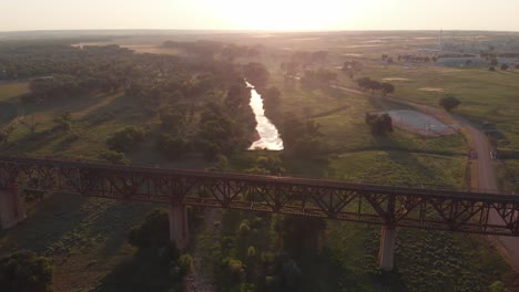 countryside scenic view of old rusty steel railroad track bridge with bright white sun on horizon, aerial rising pull back