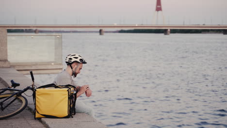 food delivery man rests next to his thermal backpack at sunset on the shore of a river and takes a look at his smartwatch to know where to go next