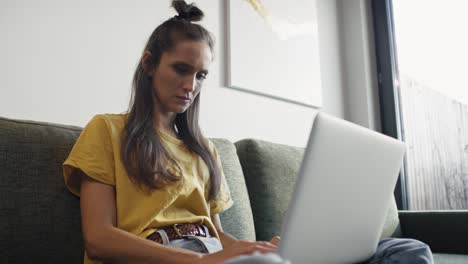 mujer caucásica seria sentada en el sofá y trabajando en la computadora