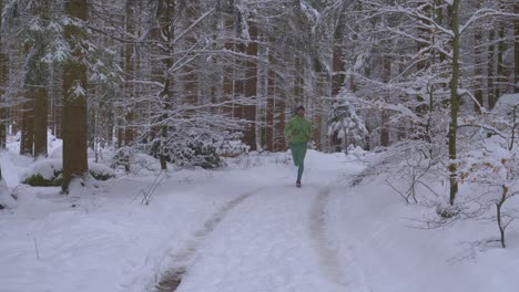 Athletischer-Erwachsener-Mann,-Der-Aus-Der-Ferne-In-Grünem-Outfit-Im-Schneebedeckten-Wald-Auf-Wildem-Pfad-Während-Des-Kalten-Wintertages-Läuft---In-Richtung-Kamera,-Zeitlupe