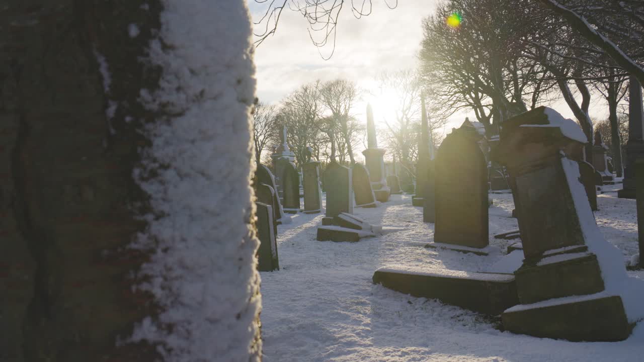 Lento movimento della camera da dietro un albero per rivelare un tranquillo cimitero a Sunderland, in Inghilterra, durante l'inverno. La neve si posa sulle lapidi inclinate sotto un cielo luminoso e nuvoloso, creando un'atmosfera fredda e riflessiva