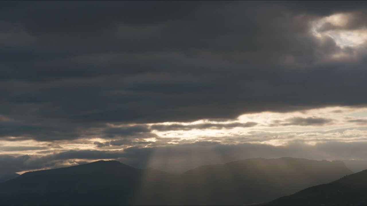 Time lapse style shot of dramatic crepuscular rays (God rays) shining through heavy clouds. Dark mountain range silhouette under an atmospheric and moody sky with natural light transitions