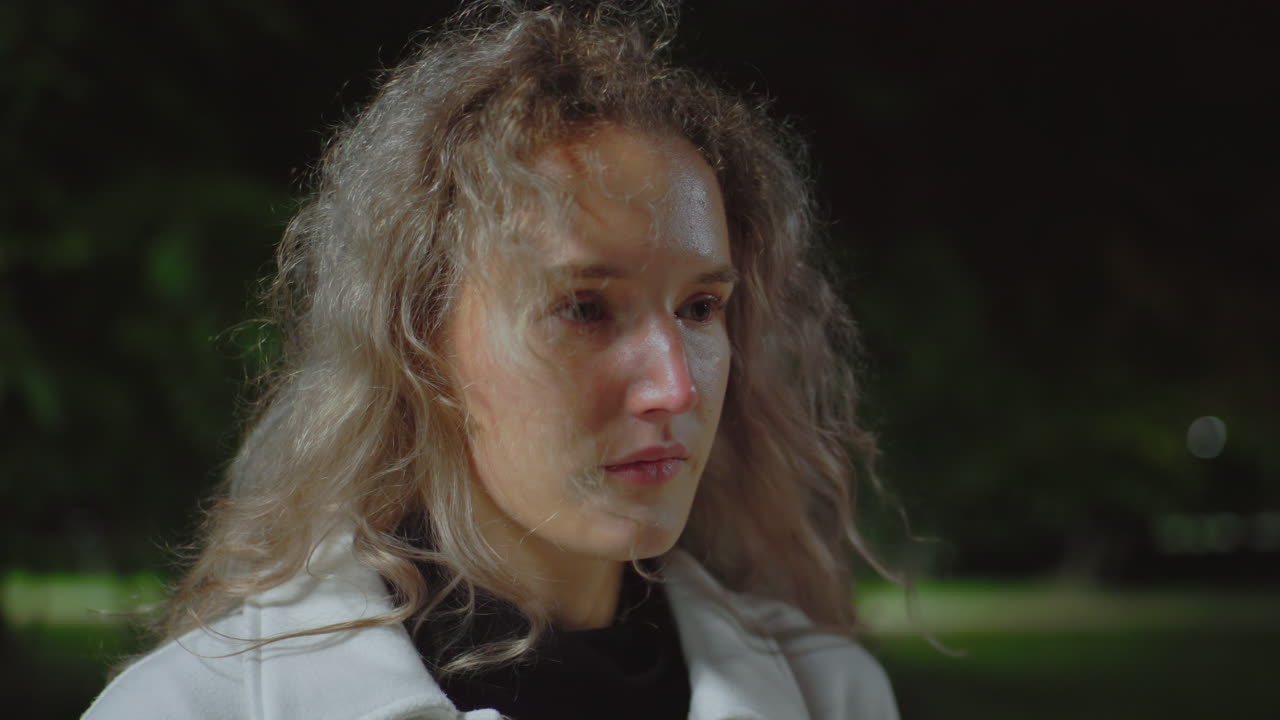 Sober lady sitting outdoors at night slowly raising head while reflecting quietly with wind gently moving her curly hair and soft night lights with blurry background of trees and dim park ambiance